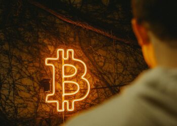 A hooded figure holds a phone displaying cryptocurrency logos against a dark urban backdrop.