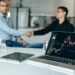 Business executives shaking hands with financial graphs and logos of Amazon and Nvidia in the background.