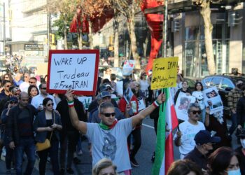Demonstrators holding signs during protests in Iran, with darkened screens in the background.