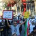 Demonstrators holding signs during protests in Iran, with darkened screens in the background.