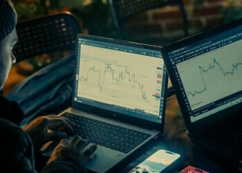 A trader monitors fluctuating cryptocurrency charts on multiple screens in a dimly lit room.