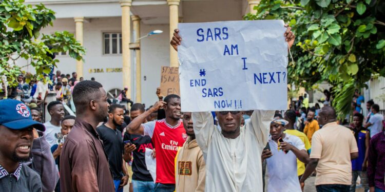 A user interacting with the Bitchat app on a smartphone amidst protest signs and unrest.