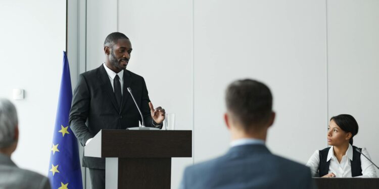 Alex Holmes speaking at a conference with Bitcoin logos and regulatory symbols in the background.
