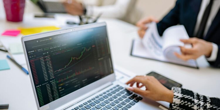 A business professional analyzes cryptocurrency charts on a laptop with financial documents nearby.