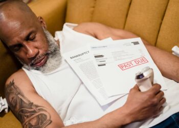 A distressed man seated at a desk, surrounded by financial documents and a laptop, depicting despair.