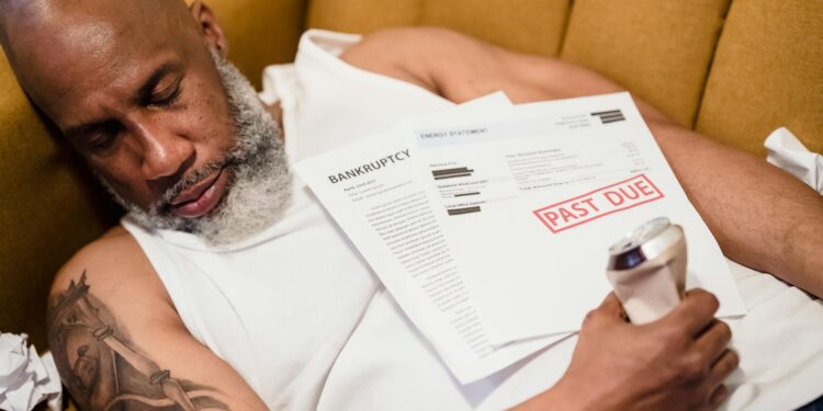 A distressed man seated at a desk, surrounded by financial documents and a laptop, depicting despair.