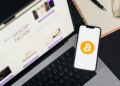 A hacker in a hoodie manipulates a laptop with Bitcoin logos on the screen.