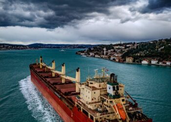 A cargo ship navigates the Strait of Hormuz with digital currency icons overlayed.