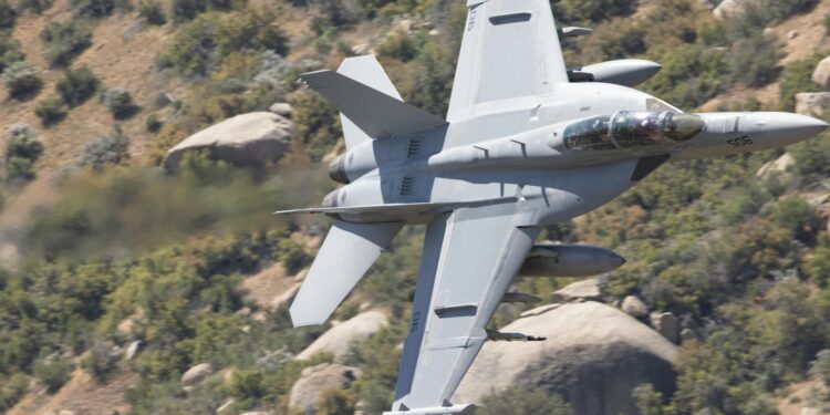 F-15E aircraft in flight over rugged terrain, with smoke rising from a distant location.