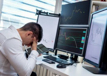 Falling stock charts and a worried trader at a financial market desk during tense economic times.