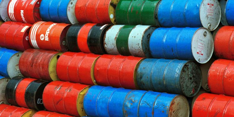 Oil barrels stacked at a port with a ship entering the Strait of Hormuz in the background.