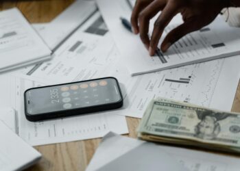 A hand holds a smartphone displaying a cryptocurrency graph and dollar bills on a table.