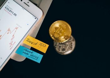 A person checks cryptocurrency prices on a smartphone with financial documents beside it.