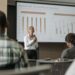 Stephanie Cutter speaking at a conference with financial graphs displayed in the background.